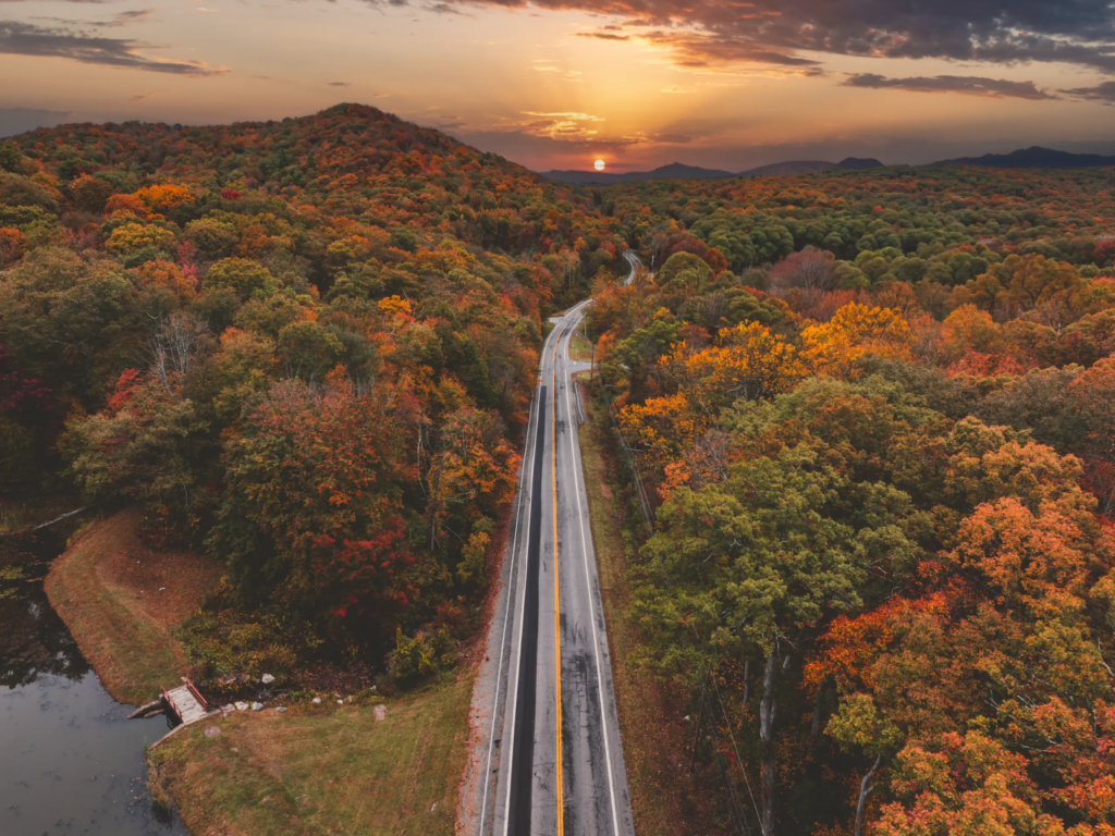 Aerial view of a two-lane highway winding through fall foliage at sunset in a mountain forest.