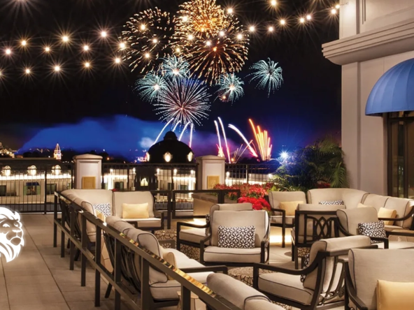 Rooftop terrace with lounge seating and string lights overlooking a colorful fireworks display at night.
