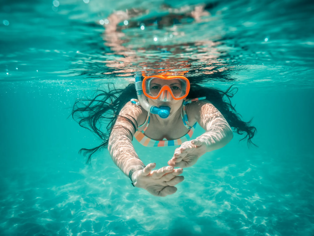 Woman snorkeling underwater in clear turquoise water, wearing an orange mask and striped swimsuit.