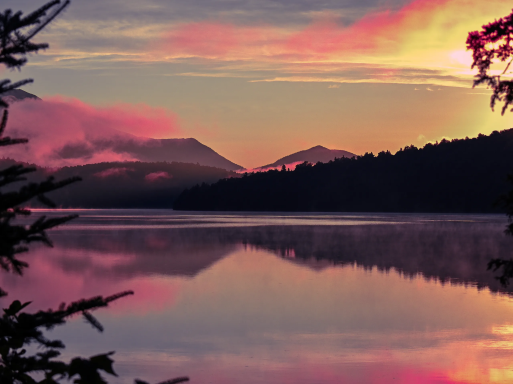 Serene mountain lake at dusk, perfect for romantic getaways in upstate NY, with pink skies and silhouetted forested hills.