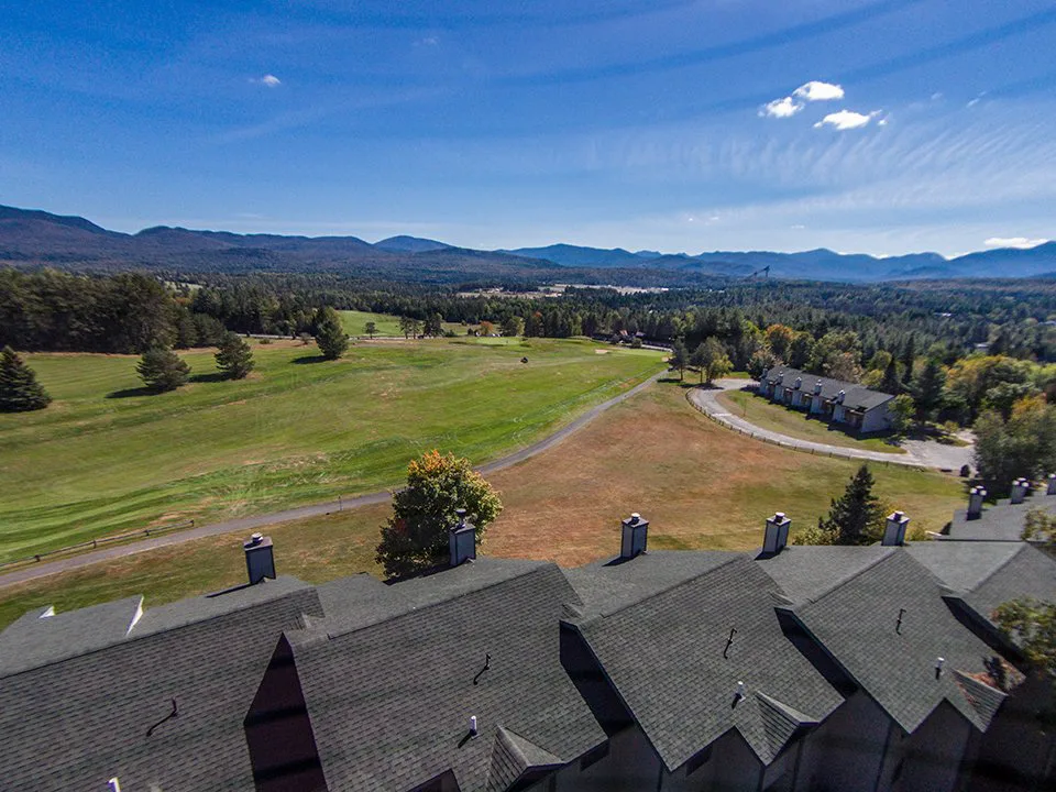 Aerial view of a resort with shingled rooftops overlooking a golf course and mountain range under a blue sky.