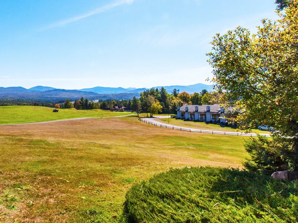 Rolling green fields with a split-rail fence, a white country house, and mountain ridgeline under a clear blue sky.