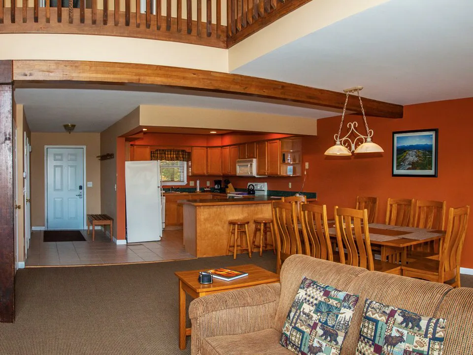 Open-plan lodge interior with wood beam accents, kitchen with bar stools, large dining table, and nature-print sofa pillows.
