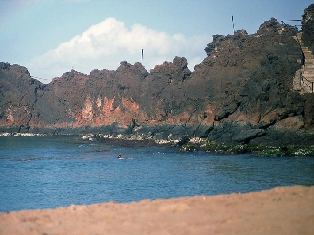 Black volcanic rock cliffs meeting the shore at Black Rock, Maui, with a swimmer visible in the calm bay.