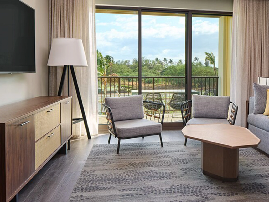 A modern living room area with gray seating and a balcony overlooking tropical trees at Hilton's Maui Bay Villas.