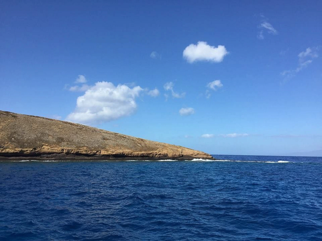 The rocky, crescent-shaped shore of Molokini Crater rising from deep blue ocean waters under a sunny, partly cloudy sky.