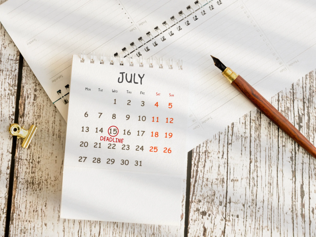 July calendar with July 15 circled in red and labeled DEADLINE, beside a fountain pen on a wood surface.