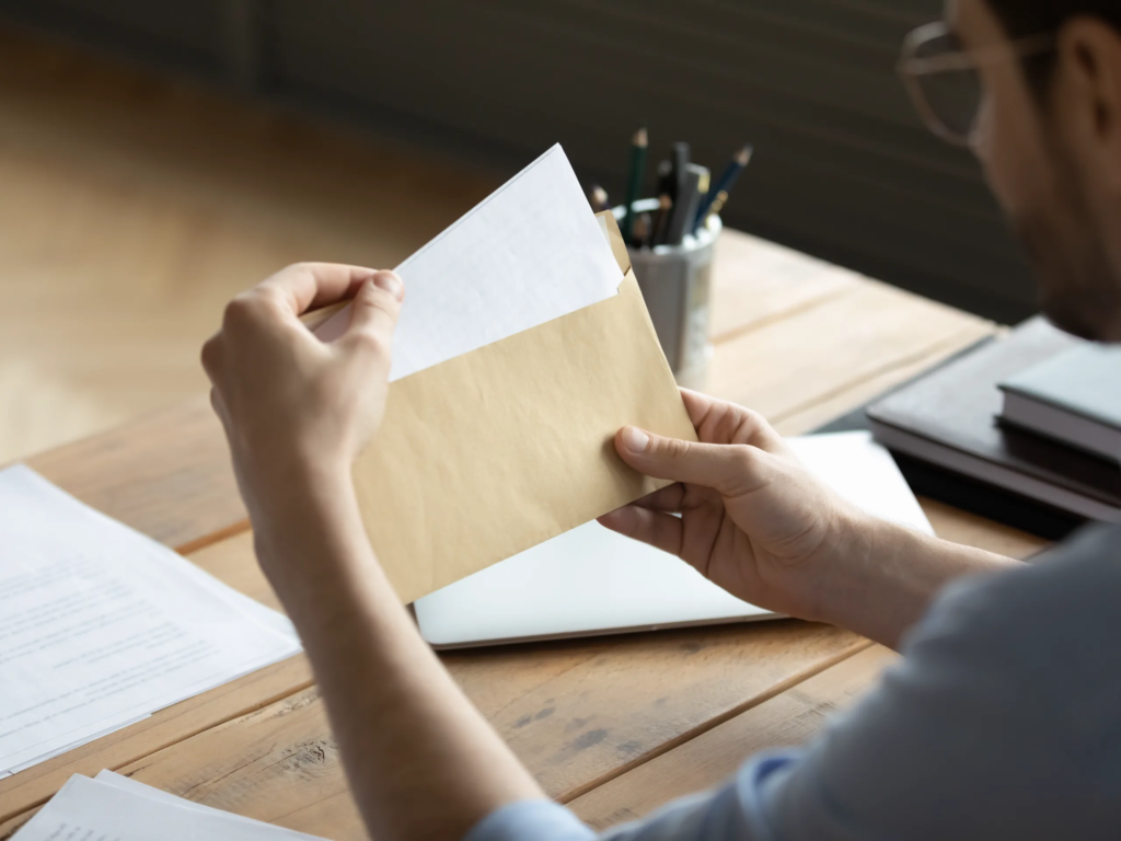Person pulling a letter from a manila envelope while sitting at a wooden desk with papers and pencils nearby.